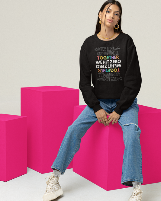 Woman sitting on bright pink cubes wearing a black crewneck sweatshirt with mirrored rainbow “TOGETHER WE HIT ZERO” graphic; part of Hit Zero Co's Beyond the Mat Collection.